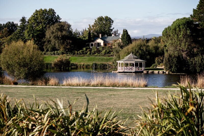 A view across a lake towards a round, lakeside pavilion and towards the heritage Cellar Door peeking through established gardens